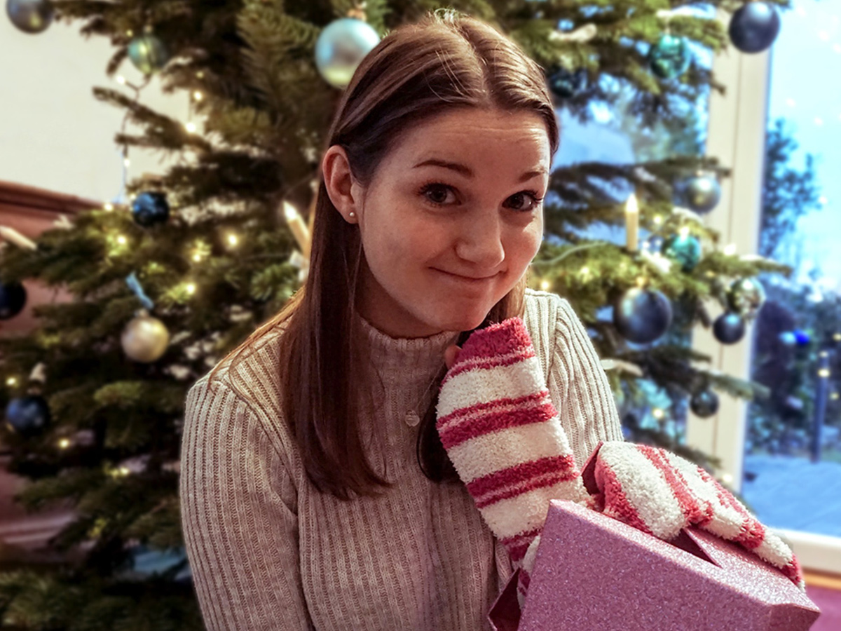 Frau mit falschem Lächeln vor einem Weihnachtsbaum mit Weihnachtsgeschenk zum Umtauschen in der Hand.
