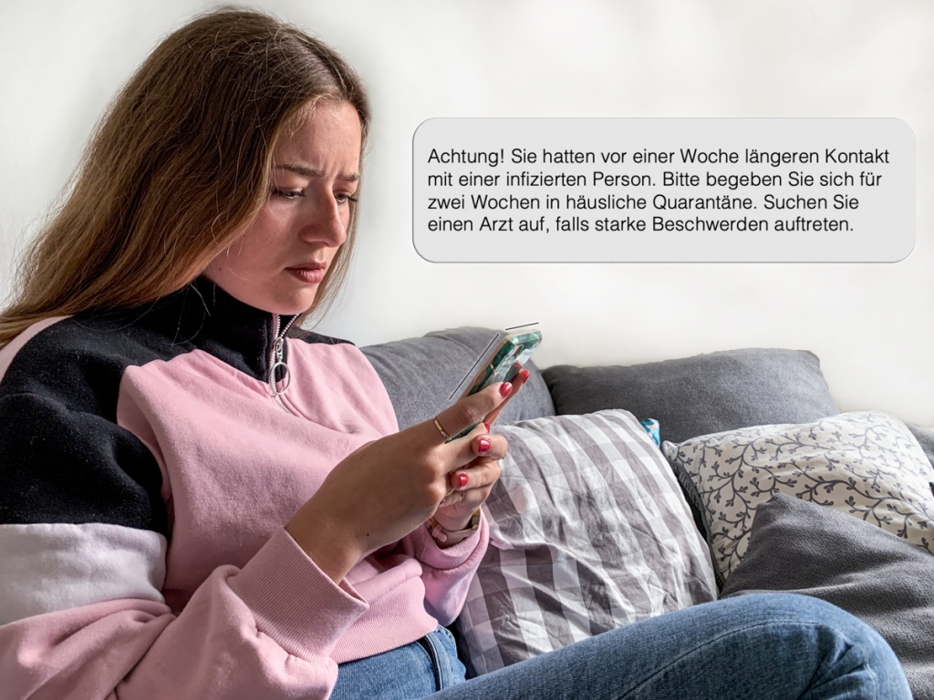 Frau auf Sofa schaut beunruhigt auf Smartphone, da Corona- Tracking-App längeren Kontakt mit infizierter Person und Aufforderung zu häuslicher Quarantäne meldet.