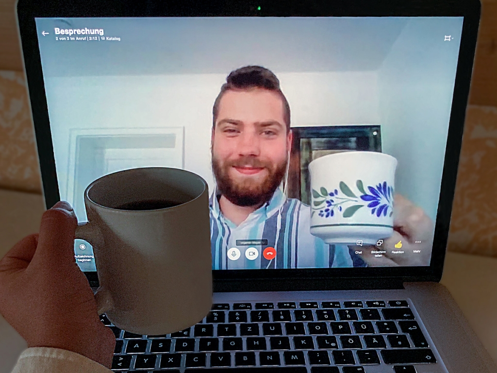 Videoanruf, bei dem sowohl Person vor dem Bildschirm, als auch Person auf dem Bildschirm Kaffeetasse im Homeoffice in der Hand hält.