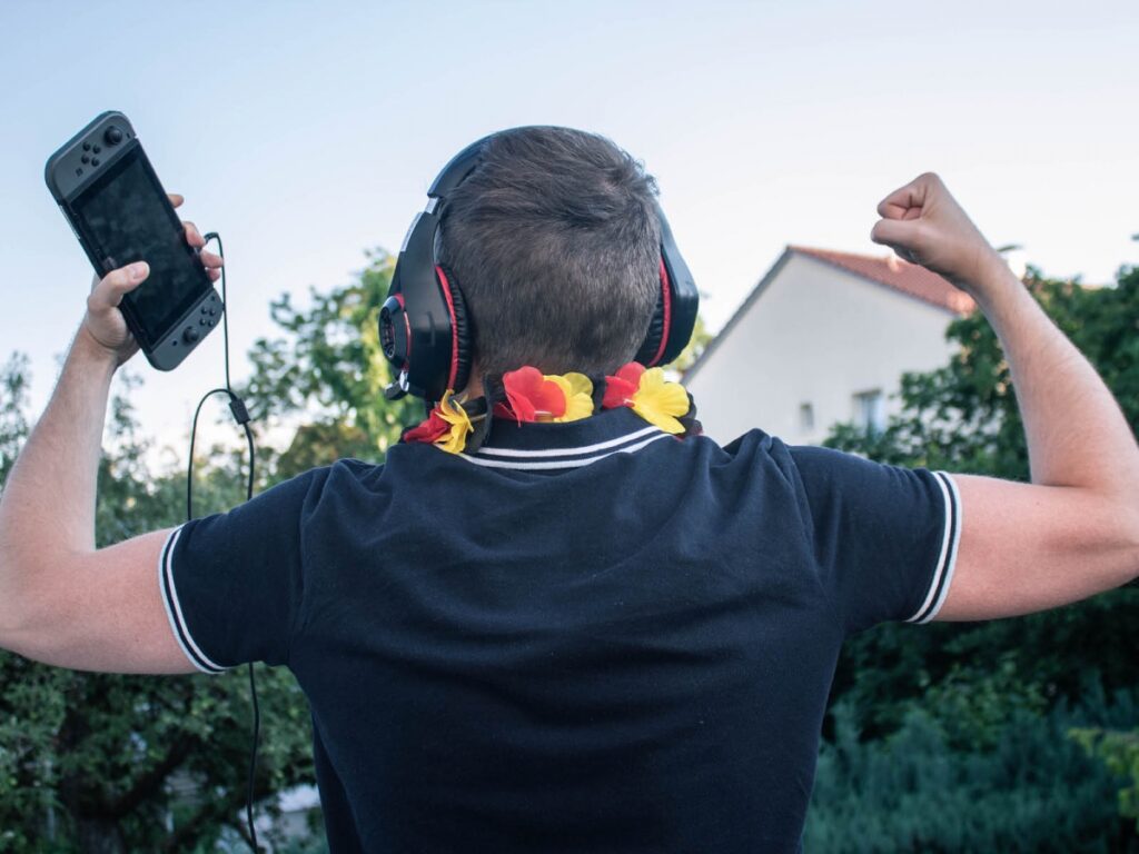 Eine Mann freut sich über seinen Sieg. Dabei trägt er eine deutsche Blumenkette, Spielekonsole und Kopfhörer, um die Games-Branche zu symbolisieren.