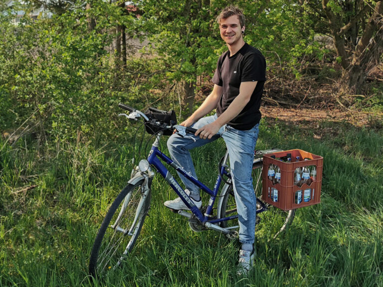 Der Start-Up Gründer sitzt auf seinem Fahrrad, auf dem Gepäckträger transportiert er eine Getränkekiste mit der Kistenhalterung.