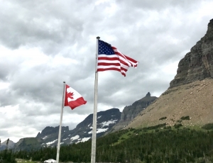 The American and Canadian flags are flying in the wind at the top of two flags poles. In the background are mountain ranges and trees.